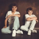 Zwei Burschen sitzen in jeans und weißen T-Shirt am boden vor einer rostigen Wand