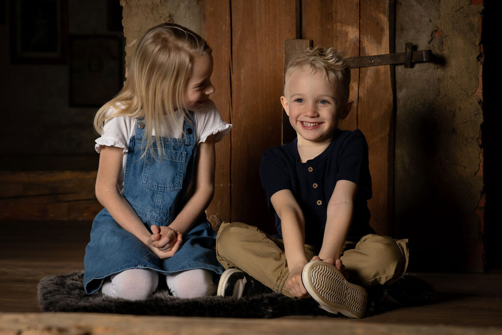 mädchen kniet am boden und lächelt den bruder an der im schneidersitz daneben sitzt und strahlt vor einer alten Selch am DachbodenFotostudio