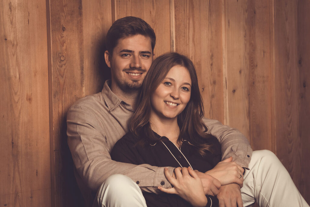 paar sitzt vor holzwand und der mann umarmt die frau liebevoll